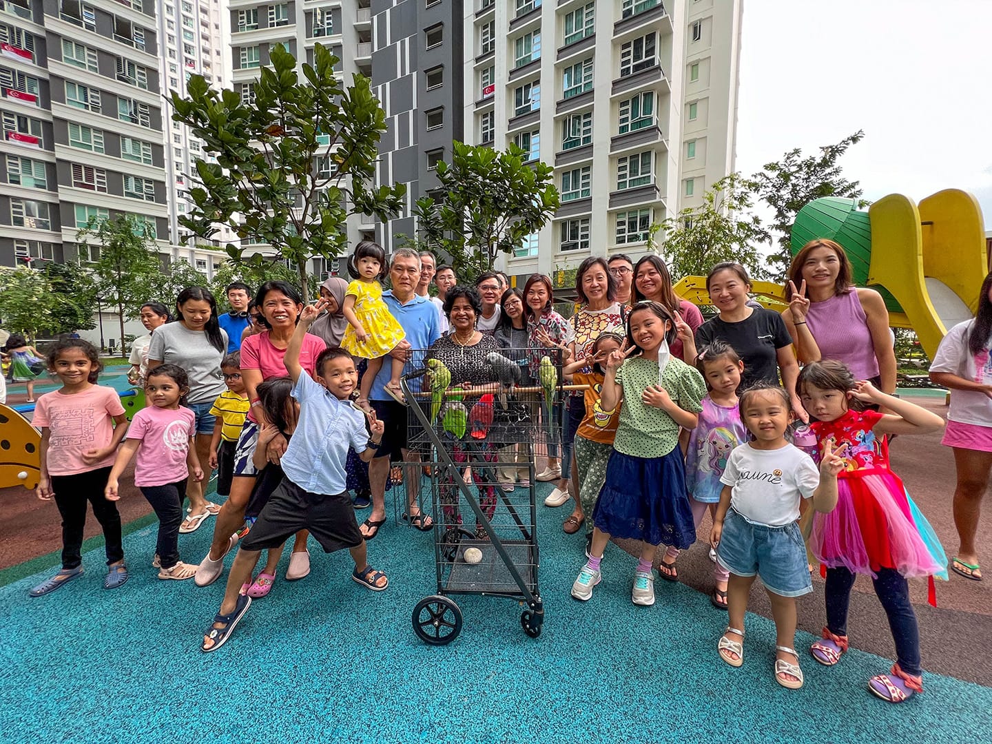 Group of adults and children pose near a wheeled cage with green parrots at an outdoor playground.